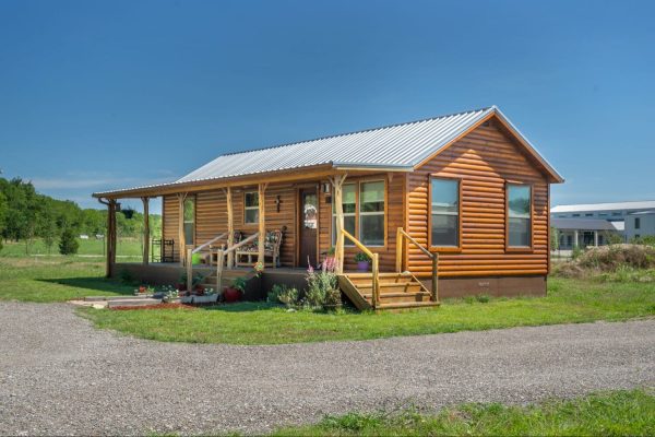 A furnished log cabin from Ulrich sits on a grass lot with a gravel path passing by it.